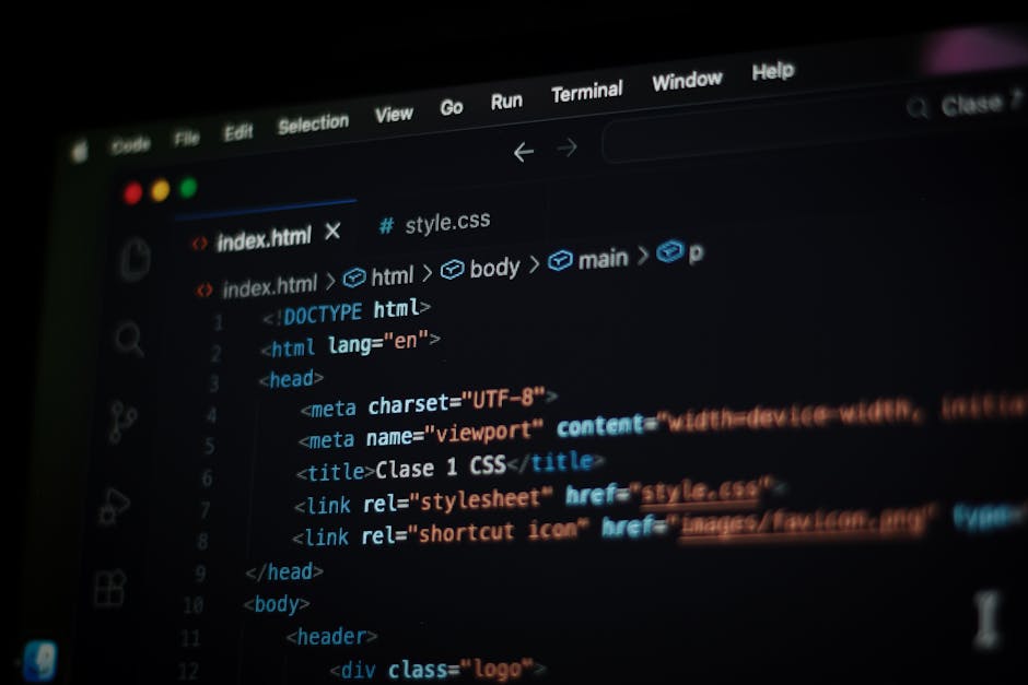 A person working on a laptop placed on a wooden desk, with their right hand resting on the keyboard. The laptop screen displays lines of coloured code on a dark background, indicating software or web
