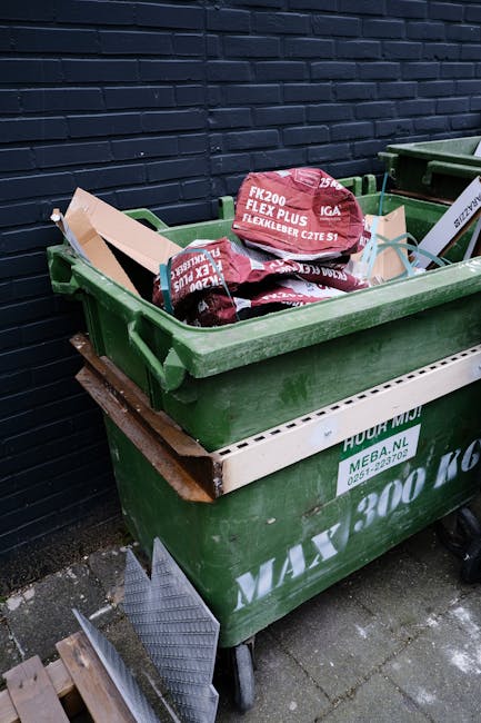 Three large black rubbish bags, made of thick plastic with a glossy finish, are positioned on the curbside pavement in front of a dark metal fence and dense foliage in the background. The bags appear 