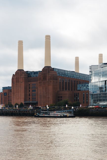 A low-angle view of a large red-brick building with multiple rectangular windows, some with metal grilles, and two tall, cylindrical beige smokestacks extending upwards against a cloudy sky. The build