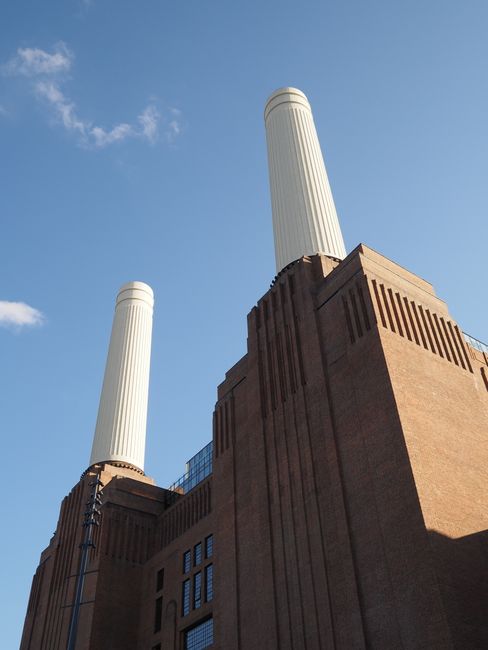 A low-angle view of a historic power station building featuring two tall, vertical, white cylindrical chimneys with ribbed textures, set against a clear blue sky with a few wispy clouds. The building'